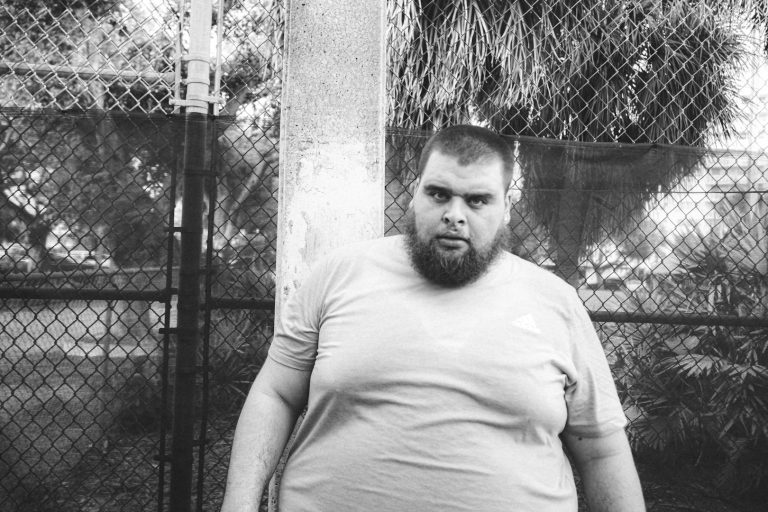 Uomo in sovrappeso con barba e maglietta grigia, in un ambiente esterno con recinzione.