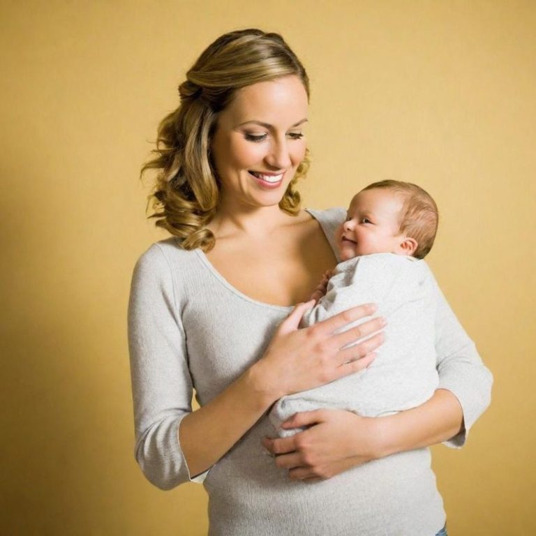 Madre sorridente tiene in braccio un neonato, entrambi vestiti in abiti chiari.
