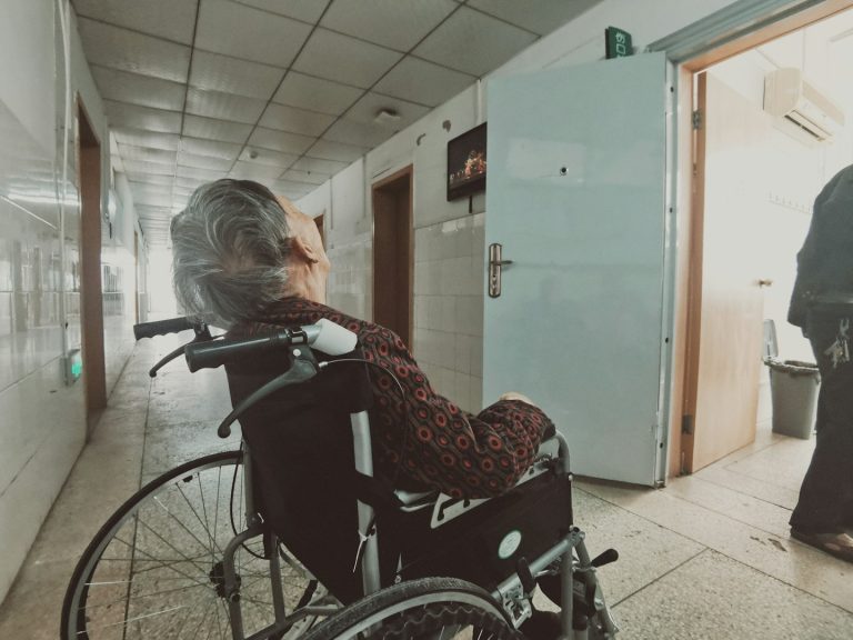An elderly person in a wheelchair looking towards an open door in a hallway.