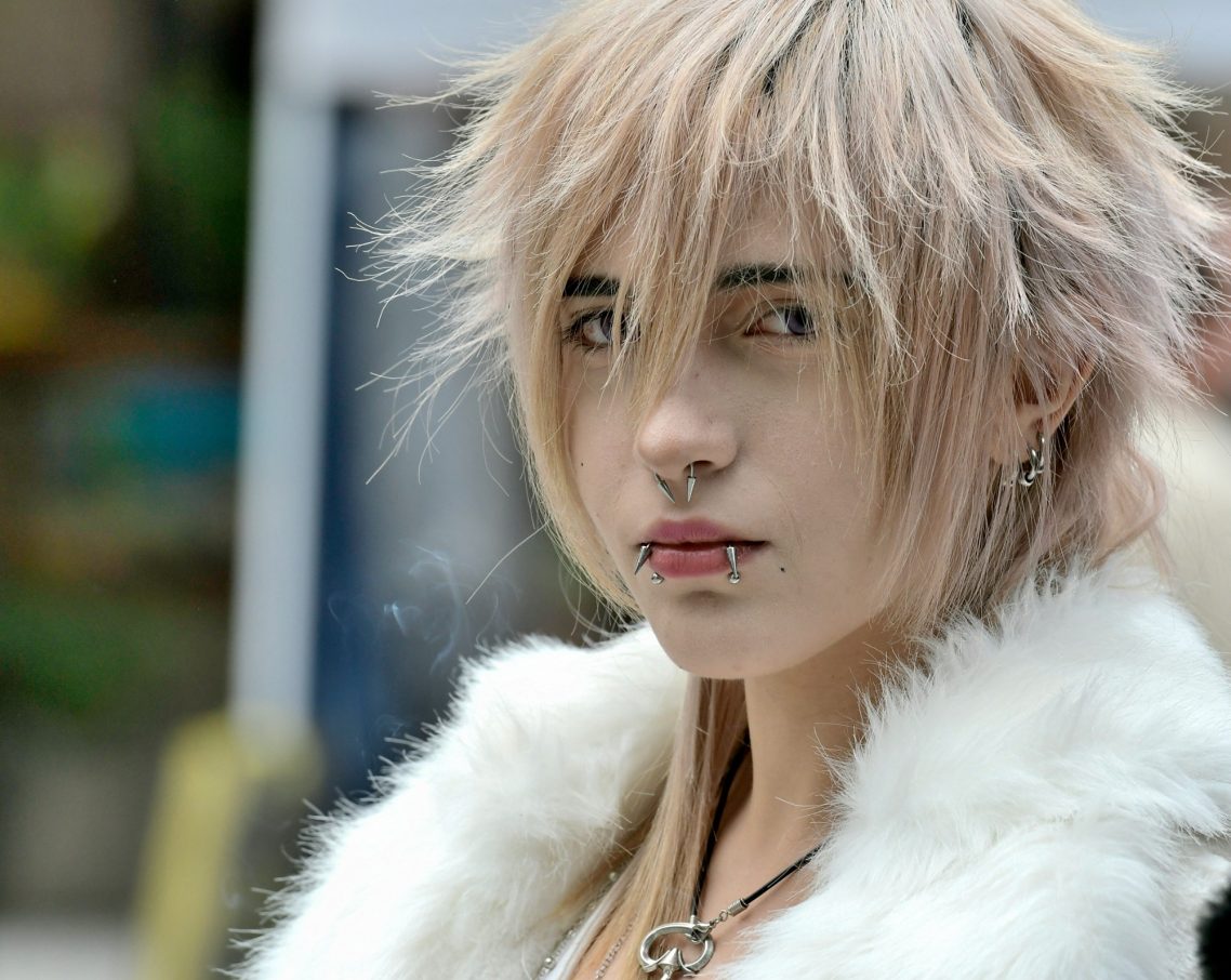 Ragazza con capelli biondi e disordinati, indossa un soprabito bianco e ha un piercing.