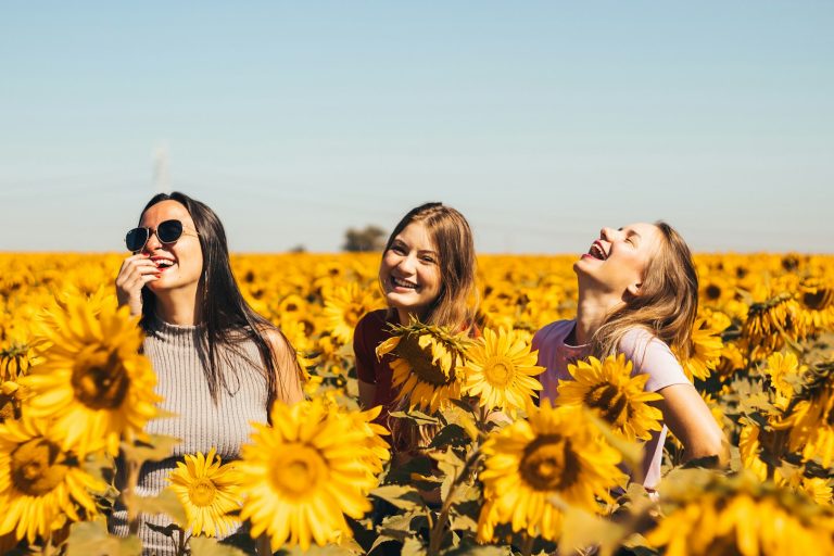 Tre donne sorridenti tra girasoli in un campo soleggiato.