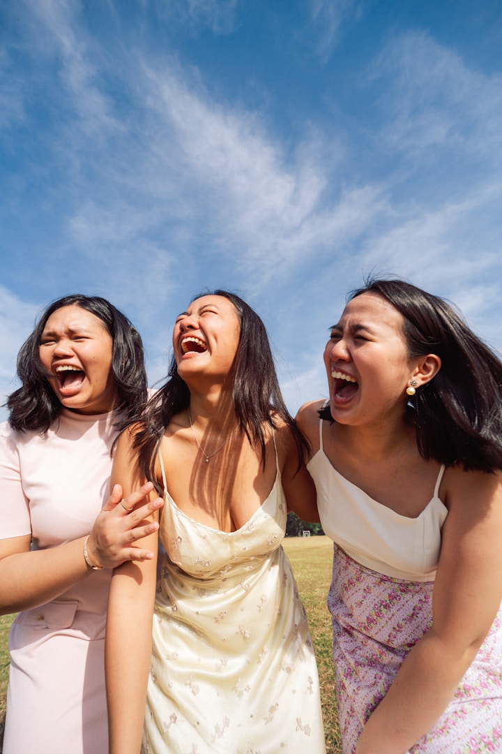 Tre giovani donne ridono felicemente insieme in un campo sotto un cielo azzurro.
