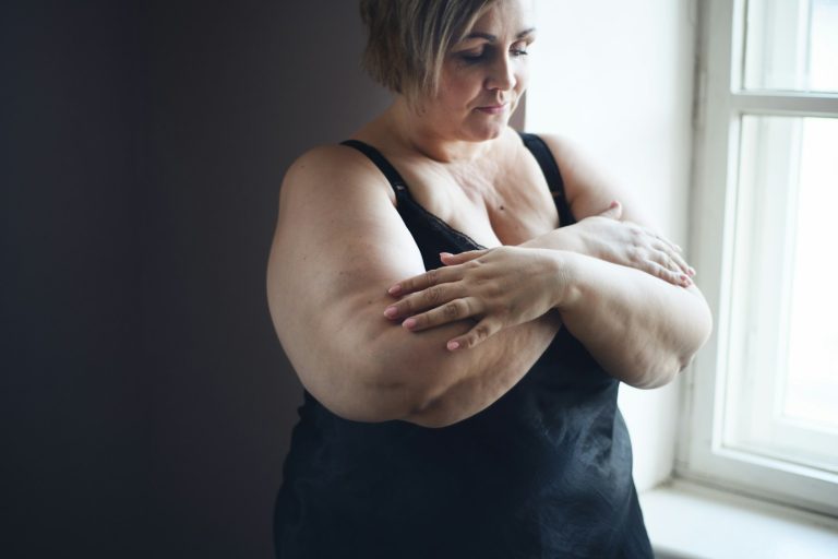 Donna obesa con capelli corti e biondi, in posa davanti a una finestra, abbigliamento nero.