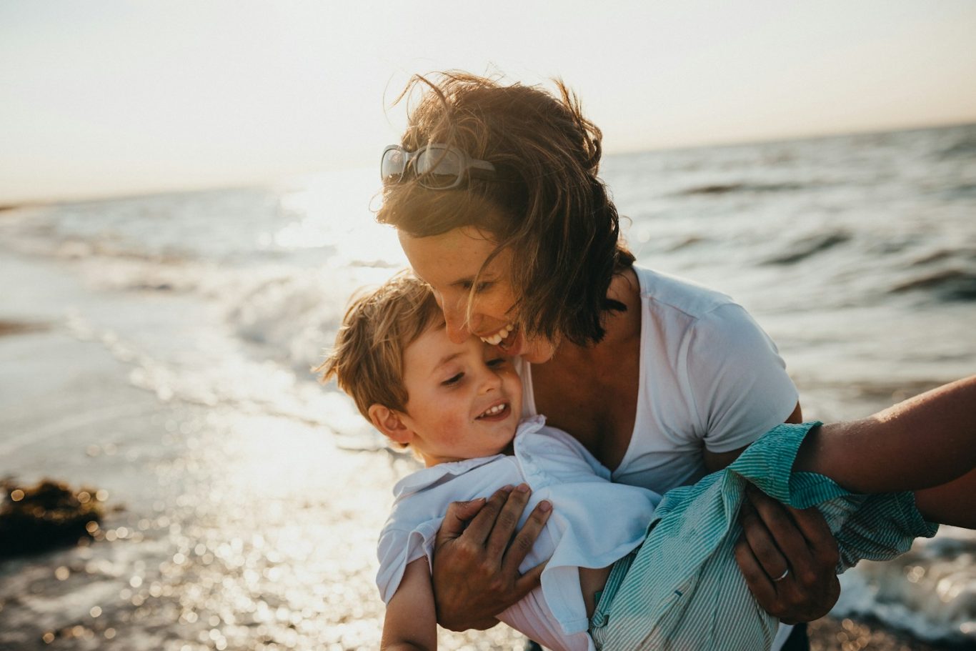 Madre tiene in braccio un bambino sorridente sulla spiaggia al tramonto.